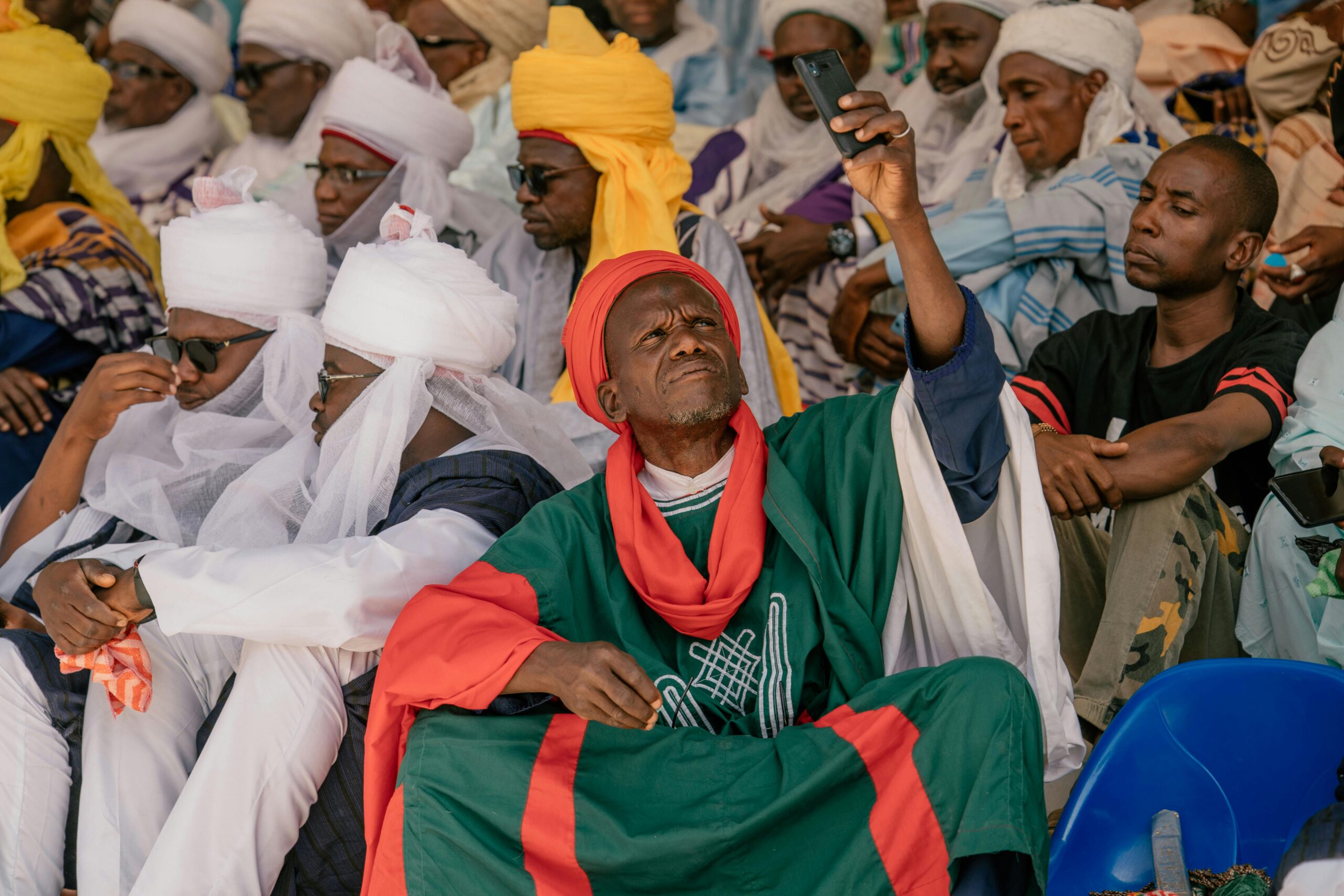 Eine Gruppe von MÃ¤nnern in traditioneller afrikanischer Kleidung hÃ¤lt bei einer Veranstaltung im Freien einen Moment mit einem Smartphone fest.
